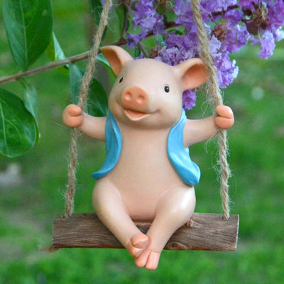Gartenfigur Schwein auf Holzschaukel mit blauer Weste, hängt an Seilen vor lila Blüten im Garten